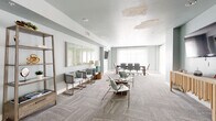 Located at 401 N Chaparral St in Corpus Christi, Texas, this single-floor multipurpose room serves as both a professional conference space and a relaxed lounge area. The open layout is unified by patterned, light-gray carpeting laid in a herringbone design, which adds visual texture and warmth throughout the space.

The conference area is anchored by a large rectangular wood-toned table surrounded by ergonomic rolling chairs, accommodating sizable group meetings. A wall-mounted flat-screen TV is positioned nearby for presentations and collaborative work. Sheer white floor-to-ceiling drapes line the exterior wall, diffusing generous natural light and contributing to the room's airy, calm atmosphere.

The lounge area features a curated mix of modern accent chairs with gold-toned frames, a small side table, and additional seating options including a warm leather chair near the entry. A decorative open shelving unit and a wooden sideboard provide functional display surfaces, while framed artwork and a table lamp add refined finishing touches.

The soft blue-gray walls create a cohesive, professional aesthetic throughout the space. Glass double doors at the entry provide easy access and a transparent connection to the adjacent corridor. Overall, the space is well-suited for meetings, community gatherings, or collaborative work sessions.