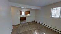 This single-floor space is designed with a practical, open layout that connects the main living area to the kitchen through a pass-through opening in a dividing wall. The living area is finished with wall-to-wall carpet and features a textured ceiling along with a baseboard heating unit positioned along the wall, providing an efficient heating solution. Natural light filters into the space, casting subtle brightness across the room.

The kitchen is accessed through the open pass-through and offers warm-toned wood cabinetry both above and below the countertops, providing ample storage. A sink is positioned beneath a window, allowing natural light into the cooking area. A range with an overhead ventilation hood is included among the kitchen appliances. The kitchen floor features a hard surface tile, contrasting with the carpeted living space and creating a clear visual distinction between the two areas.

Overall, the space presents a straightforward and functional layout with neutral wall tones throughout, making it well-suited for comfortable everyday living.