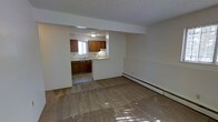 This single-floor space is designed with a practical, open layout that connects the main living area to the kitchen through a pass-through opening in a dividing wall. The living area is finished with wall-to-wall carpet and features a textured ceiling along with a baseboard heating unit positioned along the wall, providing an efficient heating solution. Natural light filters into the space, casting subtle brightness across the room.

The kitchen is accessed through the open pass-through and offers warm-toned wood cabinetry both above and below the countertops, providing ample storage. A sink is positioned beneath a window, allowing natural light into the cooking area. A range with an overhead ventilation hood is included among the kitchen appliances. The kitchen floor features a hard surface tile, contrasting with the carpeted living space and creating a clear visual distinction between the two areas.

Overall, the space presents a straightforward and functional layout with neutral wall tones throughout, making it well-suited for comfortable everyday living.