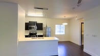 This single-floor space showcases a thoughtfully designed open-concept layout that seamlessly connects the kitchen and living areas, creating a cohesive and functional environment. The kitchen is outfitted with dark cabinetry that provides a striking contrast against the clean white countertops, including a spacious peninsula that serves as both a prep area and a casual dining or gathering spot. Stainless steel appliances, including a refrigerator, range, and over-the-range microwave, lend a polished, contemporary feel to the cooking space. Pendant lighting with sleek, modern fixtures hangs above the peninsula, complementing the overall aesthetic. The living area extends naturally from the kitchen, benefiting from a window that invites natural light into the space, enhancing its open and airy character. Rich, dark hardwood-style flooring runs throughout, grounding the neutral cream-toned walls and adding warmth and depth. An additional pendant light fixture illuminates the living area, contributing to a well-lit and welcoming ambiance. The overall design reflects a clean, modern sensibility with a cohesive palette of dark and light tones.