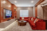Located at 837 NW Donovan Rd in Lee's Summit, Missouri, this single-floor entertainment space is thoughtfully designed to deliver a premium recreational experience. The main lounge area serves as the centerpiece, featuring a generous rust-orange velvet sectional sofa paired with a rounded wood coffee table and a large wall-mounted television framed by warm, backlit built-in wooden shelving. Textured accent walls with a subtle geometric pattern add depth and sophistication to the room, while recessed lighting creates a comfortable ambiance throughout. A sliding barn door with distinctive wooden beam framing divides zones, adding an industrial-meets-warm design aesthetic. Exposed metallic ductwork reinforces this design theme with an intentional, modern touch. A dedicated golf simulator room provides an immersive experience, complete with a projector, a turf hitting mat, and dark wood-paneled walls that enhance the focused atmosphere. Adjacent to the lounge, a shuffleboard table offers additional entertainment options within a spacious open area. Warm wall sconces illuminate a secondary seating nook, providing a more intimate setting. Patterned carpet flooring flows consistently throughout the space, tying together the various entertainment zones into a cohesive, well-appointed retreat.