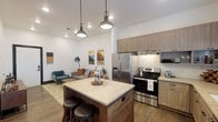 This well-designed single-floor residence blends modern aesthetics with warm, inviting elements throughout its thoughtfully arranged layout. The open-concept kitchen and living area serves as the heart of the home, where a striking wood-paneled island with light stone countertops anchors the space beneath a trio of industrial-style pendant lights. The kitchen is fully equipped with stainless steel appliances, including a French door refrigerator and electric range, complemented by light wood cabinetry, a textured tile backsplash, stone countertops, a deep undermount sink with a matte black faucet, and rustic open shelving. Wood-look flooring flows seamlessly throughout, enhancing the cohesive, contemporary feel. The living area offers a welcoming atmosphere with clean white walls, recessed lighting, and a cozy seating arrangement. Two bedrooms provide comfortable private retreats, each with soft neutral tones and natural light from well-placed windows. The bathroom features a spacious layout with dark wood vanity, stone countertops, tile flooring, and a walk-in shower with decorative tile accents. The entry includes a bold black-framed door and direct access to a secondary exterior door, adding both style and convenience to this smartly designed residence.