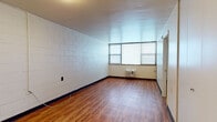 Located at 1610 N Kingshighway Blvd in St. Louis, Missouri, this single-floor unit offers a straightforward and functional layout across several distinct areas. The main living space is generously sized, showcasing warm wood-look laminate flooring and painted cinder block walls that lend the space a durable, solid character. Natural light filters in through windows fitted with blinds, and a wall-mounted air conditioning unit provides climate control. Flush-mount ceiling light fixtures illuminate the rooms efficiently. The kitchen is equipped with a black electric coil-burner range, warm wood-toned cabinetry, and granite-style laminate countertops, providing practical cooking and storage space. A separate bedroom area features its own window with a blind, continuing the wood-look flooring and cinder block wall aesthetic, along with a closet for storage. The bathroom is outfitted with a tiled shower-tub combination, a toilet, and a vanity with a rich reddish-brown cabinet, a white sink basin, and a wall-mounted mirror. Wood-look tile flooring ties the bathroom together cohesively. Throughout the unit, the consistent use of wood-look flooring and neutral painted surfaces creates a clean, utilitarian ambiance well-suited for everyday living.