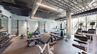 This single-floor fitness facility combines industrial design with a comprehensive range of workout amenities. The main exercise floor is an expansive, open space with exposed concrete ceilings, visible ductwork, and suspended linear lighting that lends the room a modern industrial character. Rubber flooring throughout the workout area provides durability and safety underfoot.

The facility is outfitted with an extensive selection of equipment, including Matrix-brand cardio machines such as ellipticals, treadmills, and stationary bikes, as well as a full complement of strength training equipment—power racks, adjustable benches, cable machines, free weights, and kettlebells. Mirrored walls along one side enhance the sense of space, while a row of floor-to-ceiling windows along the perimeter floods the workout floor with natural light and offers views of the surrounding outdoor landscape.

A dedicated wellness area features a glass-enclosed infrared sauna with wood-paneled interior, accompanied by an adjacent room with direct access to an outdoor terrace. The support areas include a well-maintained locker corridor with tile flooring, private changing rooms, water stations, and recessed lighting, providing a clean and functional transition space for users entering and exiting the facility.