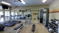 This fitness center is a fully equipped exercise facility designed to support a wide range of workout routines. The space features a thoughtfully arranged layout that accommodates both cardiovascular and strength training needs. Along one wall, a row of cardio machines — including treadmills and ellipticals — provides ample options for aerobic exercise. The strength training area is anchored by a multi-function cable machine and a power tower for bodyweight exercises, complemented by weight benches suitable for a variety of lifts. Large mirrors line the walls, enhancing the sense of openness while allowing users to monitor their form during workouts. The flooring consists of durable, cushioned carpet tiles well-suited for a fitness environment. Overhead, ceiling fans and bright lighting ensure the space remains comfortable and well-illuminated during use. The neutral-toned walls with a contrasting accent border add a clean, functional aesthetic to the room. A vaulted ceiling contributes to an airy, open atmosphere throughout the space.