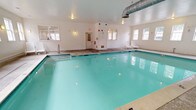 This indoor pool facility offers a clean, well-maintained aquatic environment designed for year-round use. The centerpiece is a generously proportioned swimming pool with clear turquoise water, illuminated by underwater lighting that enhances visibility and ambiance. Entry into the pool is facilitated by stainless steel handrails and built-in steps, ensuring safe and convenient access for all users.

The pool deck is finished in neutral-toned stone tile, providing a slip-resistant surface that surrounds the pool on multiple sides. White lounge chairs are arranged along the perimeter, offering comfortable poolside seating. The walls are painted in a crisp, bright white, contributing to the space's clean and airy atmosphere.

Natural light is introduced through numerous multi-pane windows lining multiple walls, providing views of the surrounding exterior while keeping the space feeling open and well-lit. Wall-mounted sconce lighting supplements natural light throughout the room. Visible HVAC ductwork along the ceiling ensures proper climate control and ventilation.

Safety equipment, including a life ring and emergency board, is mounted on the wall alongside clearly marked exit signage and fire safety devices. A dedicated entrance door with sidelights provides direct access to the pool area from outside.