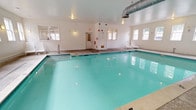 This indoor pool facility offers a clean, well-maintained aquatic environment designed for year-round use. The centerpiece is a generously proportioned swimming pool with clear turquoise water, illuminated by underwater lighting that enhances visibility and ambiance. Entry into the pool is facilitated by stainless steel handrails and built-in steps, ensuring safe and convenient access for all users.

The pool deck is finished in neutral-toned stone tile, providing a slip-resistant surface that surrounds the pool on multiple sides. White lounge chairs are arranged along the perimeter, offering comfortable poolside seating. The walls are painted in a crisp, bright white, contributing to the space's clean and airy atmosphere.

Natural light is introduced through numerous multi-pane windows lining multiple walls, providing views of the surrounding exterior while keeping the space feeling open and well-lit. Wall-mounted sconce lighting supplements natural light throughout the room. Visible HVAC ductwork along the ceiling ensures proper climate control and ventilation.

Safety equipment, including a life ring and emergency board, is mounted on the wall alongside clearly marked exit signage and fire safety devices. A dedicated entrance door with sidelights provides direct access to the pool area from outside.