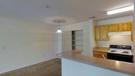 Located at 4545 Cary Glen Blvd in Cary, North Carolina, this single-floor residence offers a thoughtfully designed open-concept layout that seamlessly connects the kitchen and living areas. The kitchen is equipped with warm-toned wooden cabinetry, a countertop workspace, and a peninsula bar that extends into the living space, providing both additional prep area and a casual dining spot. A decorative pendant light hangs above the peninsula, adding a focused accent to the open layout. The kitchen also features a built-in sink and overhead ventilation, reflecting practical design choices throughout. Adjacent to the kitchen, a built-in shelving unit offers generous storage and display space, ideal for pantry items or organizational use. The living area is finished with plush carpeting that adds warmth and comfort underfoot. Neutral white walls and standard-height ceilings keep the interior bright and versatile, allowing for a variety of furnishing styles. Ceiling vents ensure consistent climate control throughout the space. Overall, the layout promotes an easy flow between functional zones, making it a practical and comfortable single-level home.