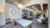 This well-appointed clubroom offers a dynamic, multi-functional gathering space designed for both leisure and socializing. At the heart of the room sits a full-size billiards table, positioned beneath a striking cluster chandelier composed of globe-shaped lights that serves as a bold focal point. The open-concept layout flows seamlessly into a wet bar area, outfitted with a mounted television, backsplash tiling, and ample counter space — ideal for entertaining. Adjacent to the bar, a high-top dining table with bar seating provides a casual spot for conversation or dining. Toward the back of the space, a relaxed lounge area is furnished with comfortable seating and accented by warm, ambient lighting, creating an inviting retreat. The ceiling design features exposed dark wood beams alongside recessed and pendant lighting throughout, adding architectural depth and visual interest. Contemporary artwork adorns the walls, including a large-scale diamond print that lends a modern, curated aesthetic. Neutral tones in the carpeting and walls are complemented by teal and warm wood accents, creating a cohesive and sophisticated atmosphere throughout the clubroom.