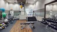 Located on E Old River Rd in Jacksonville, FL, this single-floor fitness center offers a well-organized and comprehensive workout facility. The space is divided into two distinct zones connected by a short staircase with a railing, providing clear separation between cardio and strength training areas.

The cardio room features light tile flooring, sage-green walls, and large windows that flood the space with natural light and offer direct views of the outdoor pool area. Multiple treadmills and cardio machines are arranged to take full advantage of these outdoor views. Recessed lighting and a textured ceiling complete the bright, open atmosphere.

The strength training room is outfitted with wood-style flooring and rubber matting, housing a cable machine, adjustable weight bench, a full dumbbell rack with mirror backing, resistance accessories, stability balls, and exercise mats. Ceiling fans, recessed lighting, and a wall-mounted TV contribute to a functional and energetic environment. Striped wall detailing adds a modern design touch throughout.

Adjacent to the cardio area is a kitchenette featuring dark-toned cabinetry, granite-style countertops, stainless steel appliances, and bar-height seating — providing a convenient refreshment space. Framed artwork is displayed throughout, adding a polished, community-oriented aesthetic to the overall facility.