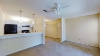 This single-floor space is designed with an open-concept layout that connects the kitchen and living area, creating a cohesive and functional flow. The living area is finished with wall-to-wall carpet in a neutral beige tone, contributing to a warm and understated ambiance. A ceiling fan with a light fixture is centrally mounted overhead, providing both air circulation and ambient lighting.

The kitchen is positioned adjacent to the living space, separated by a raised breakfast bar that doubles as a countertop divider. White cabinetry lines the kitchen walls, complemented by a black range and pendant lighting that hangs above the bar area, adding a subtle decorative touch. The kitchen features an overhead vent hood integrated beneath the upper cabinets.

A structural column serves as a natural visual divider between the living area and a secondary carpeted space, which leads toward the entry door. The entryway is straightforward and unobstructed, offering a clean transition into the interior. The walls throughout are painted in a soft, neutral cream, and standard recessed vents are visible in the ceiling. Overall, the space presents a clean, functional, and move-in ready environment.