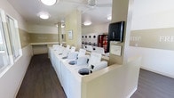 This single-floor laundry facility is designed for efficiency and ease of use, offering a clean and organized environment for all laundry needs. The room is laid out with a generous row of top-load washing machines along a central dividing counter, providing ample capacity for multiple users at once. A bank of stacked dryer units lines the back wall, maximizing the use of vertical space and accommodating high-volume laundry loads.

The interior features a neutral, warm color palette with cream and tan tones on the walls, complemented by dark hardwood-style flooring that adds a touch of warmth to the utilitarian space. Helpful word prompts — "Fold," "Fluff," and "Rise" — are displayed along the walls, adding both a decorative and instructional element to the room.

Overhead, flush-mount ceiling lights and a ceiling fan ensure the space remains well-lit and comfortably ventilated throughout the day. A paper towel dispenser and informational signage are conveniently mounted near the machines for user reference. A vending machine is also available, offering added convenience for patrons. The overall ambiance is clean, functional, and thoughtfully arranged to support a smooth and practical laundry experience.