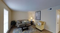 This single-floor residence offers a comfortable and well-appointed living space designed with a clean, neutral aesthetic. The living area serves as the focal point of the home, featuring smooth, light-colored walls and a flat ceiling that contribute to a bright and airy atmosphere. The space is furnished with a classic sofa accented by blue floral throw pillows, complemented by a plush yellow armchair that adds a warm pop of color. A dark wood coffee table anchors the seating arrangement atop a soft blue and yellow botanical-patterned area rug, adding texture and visual interest to the room. A matching end table with a warm lamp provides ambient lighting, creating an inviting glow. A framed figurative artwork adorns one wall, serving as a stylish focal point. Lush indoor greenery in the corner adds a touch of nature to the interior. The neutral tile flooring runs throughout, providing a cohesive and easy-to-maintain foundation. Overall, the space strikes a balance between functional design and understated elegance, making it a comfortable and inviting environment suited for everyday living.