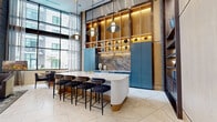 This elegantly designed single-floor space makes a striking impression through its dramatic double-height ceilings and expansive floor-to-ceiling windows, which bathe the interior in abundant natural light while offering views of the surrounding outdoors. Large-format marble-look porcelain tile flooring flows seamlessly throughout, anchoring the refined aesthetic.

The centerpiece of the space is a beautifully appointed wet bar and kitchen island area featuring a fluted white stone island with brass fixtures, surrounded by velvet-upholstered barstools with gold accents. Deep blue custom cabinetry lines the back wall, accented by a dramatic marble slab backsplash, warm wood shelving with integrated lighting, and a curated display of decorative geode accents overhead.

The lounge area is anchored by a bold grey marble fireplace surround with a linear flame feature, complemented by an arrangement of contemporary seating including leather sofas, upholstered chairs, and a patterned area rug with geometric motifs. Sculptural metal wall art adds an artistic focal point above the seating.

Additional details include illuminated built-in dark wood bookshelves, exposed grey brick columns, and a console with media display. The overall ambiance is one of upscale, contemporary luxury — sophisticated yet welcoming.