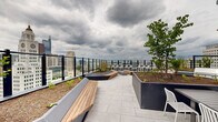 This expansive rooftop terrace is a thoughtfully designed outdoor amenity space set high above the urban landscape, offering panoramic views of a dynamic city skyline. The open-air deck is finished with large-format light grey stone pavers that lend a clean, contemporary aesthetic throughout.

Geometric raised planters, clad in dark metal paneling, are integrated throughout the terrace and filled with young trees, shrubs, and greenery, providing a natural contrast to the urban surroundings. Warm-toned hardwood benches with sleek metal accents are arranged throughout the space, offering ample casual seating in multiple zones.

A dedicated outdoor kitchen and dining area features a row of stainless steel grills set against a large-format tiled wall, illuminated by striking copper pendant lights. A slatted outdoor dining table surrounded by white modern chairs provides a comfortable space for al fresco meals.

A standout feature of the terrace is a set of suspended wooden swings mounted from a bold black steel frame, positioned to capture unobstructed views of the city below. Glass panel railings run the perimeter, ensuring safety while preserving the sweeping sightlines to the surrounding skyline. The blue and grey architectural cladding of the rooftop structure adds a modern, bold visual anchor to the space. Overall, the terrace balances functionality, greenery, and design to create a versatile and inviting outdoor retreat.