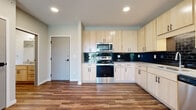 This single-floor unit presents a thoughtfully designed layout with a contemporary aesthetic throughout. The kitchen serves as a standout feature, outfitted with light natural wood cabinetry paired with sleek black hardware, a bold dark subway tile backsplash, stainless steel appliances including a range, microwave, dishwasher, and refrigerator, and light-toned countertops. Rich hardwood-style vinyl flooring runs through the kitchen and main living areas, adding warmth and durability.

The spacious living area features exposed silver ductwork that lends an industrial accent to the otherwise clean, neutral interior. Sliding glass doors lead to a private balcony with outdoor views of greenery, filling the space with natural light. The floor plan, visible in the 3D model, reveals multiple rooms connected by a practical corridor with barn-style sliding doors adding architectural interest.

Bedrooms are finished with plush carpeting and neutral-toned walls, providing a comfortable contrast to the harder surfaces in the main living areas. The bathroom includes hardwood-style flooring, a light wood vanity, and a tub-shower combination. Recessed lighting throughout maintains a clean, modern ambiance across the entire unit.
