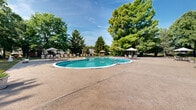 Located at 305 Deer Creek Dr in Plainsboro, NJ, this single-story community amenity facility centers around a generously sized freeform swimming pool with a classic curved design and brick-trimmed edges. The pool area is complemented by a broad concrete sundeck furnished with lounge chairs, umbrella-shaded seating groups, and ample open space for relaxation and recreation. The surrounding grounds are extensively landscaped, featuring mature deciduous and evergreen trees that provide natural shade and a sense of seclusion. Well-maintained lawns, trimmed hedgerows, and carefully arranged plantings enhance the park-like atmosphere throughout the property. A black wrought-iron perimeter fence defines and secures the pool area while preserving open sightlines to the greenery beyond. The clubhouse building presents a charming architectural design with cedar shake siding, a pitched gabled roof, stone accents at the base, and large multi-pane windows that bring in natural light. A wide concrete apron in front of the building provides convenient access and additional gathering space. Together, the pool, sundeck, clubhouse, and surrounding open lawn areas create a well-appointed outdoor recreational environment ideal for community use.