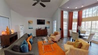 This single-story leasing office is designed to create a warm and professional first impression. The space is characterized by soaring vaulted ceilings that amplify the sense of openness, complemented by rich hardwood-style flooring throughout. Recessed lighting and a ceiling fan provide both functionality and comfort across the main area.

The lounge area serves as the focal point of the office, furnished with a plush grey sectional sofa accented by teal and orange throw pillows, patterned accent chairs, and a glass-top coffee table set atop a vibrant orange area rug. A wall-mounted flat-screen television and a wooden credenza with a coffee station complete this welcoming gathering space.

Natural light flows generously through multiple large windows dressed with white blinds and striking burnt-orange drape panels, offering views of the surrounding greenery. A dedicated round dining table with upholstered chairs nestles beside these tall windows, ideal for private consultations.

The workspace area features a substantial wooden desk with seating for staff and visitors, positioned near additional windows that further enhance the bright, airy atmosphere. A hallway with tile flooring leads to additional rooms at the rear, accessible through a glass-panel door, rounding out this thoughtfully appointed leasing office.