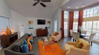 This single-story leasing office is designed to create a warm and professional first impression. The space is characterized by soaring vaulted ceilings that amplify the sense of openness, complemented by rich hardwood-style flooring throughout. Recessed lighting and a ceiling fan provide both functionality and comfort across the main area.

The lounge area serves as the focal point of the office, furnished with a plush grey sectional sofa accented by teal and orange throw pillows, patterned accent chairs, and a glass-top coffee table set atop a vibrant orange area rug. A wall-mounted flat-screen television and a wooden credenza with a coffee station complete this welcoming gathering space.

Natural light flows generously through multiple large windows dressed with white blinds and striking burnt-orange drape panels, offering views of the surrounding greenery. A dedicated round dining table with upholstered chairs nestles beside these tall windows, ideal for private consultations.

The workspace area features a substantial wooden desk with seating for staff and visitors, positioned near additional windows that further enhance the bright, airy atmosphere. A hallway with tile flooring leads to additional rooms at the rear, accessible through a glass-panel door, rounding out this thoughtfully appointed leasing office.