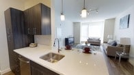 This 2-bedroom, 2-bathroom residence is designed with a modern, open-concept layout that promotes a bright and airy atmosphere throughout. The kitchen serves as a central hub, featuring a sleek quartz countertop peninsula with an undermount stainless steel sink and stylish pendant lighting that illuminates the space with a warm glow. Rich, dark-toned cabinetry provides ample storage while adding a striking contrast against the light countertops and neutral walls. The kitchen flows seamlessly into the living area, where high ceilings amplify the sense of openness and a large window invites an abundance of natural light. Track lighting overhead complements the pendant fixtures, ensuring the space is well-lit throughout the day and evening. The living area offers comfortable room for entertaining or relaxing, with hardwood-style flooring underfoot adding warmth and continuity across the open floor plan. The overall ambiance is clean, contemporary, and welcoming, with a thoughtful combination of finishes and fixtures that balance both form and function throughout this well-appointed single-floor residence.