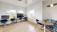 Located at View at Starr Pass in Tucson, Arizona, this single-room office delivers a clean, modern aesthetic designed to support a productive and welcoming work environment. The space is finished with light wood-style flooring throughout, lending warmth and continuity to the interior. Crisp white walls are complemented by a striking branded feature wall bearing the "View at Starr Pass" logo, rendered in bold dimensional lettering over a stylized mountain mural — serving as a clear focal point and identity anchor for the space. Multiple square windows are positioned along the walls, drawing in generous natural light and offering views of the surrounding Tucson landscape, including desert plantings and neighborhood scenery. A ceiling fan with integrated lighting provides both airflow and illumination, supporting comfort in the Arizona climate. The office is thoughtfully arranged with a workstation desk equipped with monitors, rolling chairs, and a small round meeting table with seating — accommodating both focused work and informal collaboration. A frosted-glass interior door connects to an adjacent area, adding privacy without sacrificing light. Framed artwork and Tucson-themed prints on one wall contribute a locally inspired character to the overall design. The result is a functional, well-lit, and professionally presented office space.