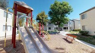 Located at 3030 W Acacia Ave in Hemet, California, this residential community is set across thoughtfully landscaped grounds that foster a welcoming neighborhood atmosphere. The property features multiple two-story buildings with neutral stucco exteriors, exterior stairways, and private balconies that contribute to a classic California residential aesthetic. The outdoor common areas are a standout feature of this community. A dedicated children's playground is equipped with a multi-slide structure, climbing elements, and colorful play equipment set over a wood chip safety surface, accompanied by nearby benches for seating. Mature trees throughout the property provide generous shade and a sense of established greenery, enhancing the overall outdoor environment. Open grassy areas between the buildings offer additional communal space, bordered by concrete pathways and lamp posts that ensure accessibility and visibility throughout the grounds. Landscaping includes a mix of shrubs, mulched beds, and maintained lawn areas. Central air conditioning units are present at each building, reflecting practical residential infrastructure. The community's layout encourages outdoor activity and neighborly interaction, making it a functional and livable environment in the Hemet area of Southern California.