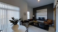 This single-floor space showcases a thoughtfully designed open-concept layout that integrates a living area and dining space into one cohesive environment. The living area is anchored by a striking dark accent wall adorned with a large-scale abstract artwork in warm earth tones of gold, beige, and charcoal, providing a bold focal point. A flat-screen television is mounted on a sleek media console beneath the artwork, complemented by a floor lamp and a plush sectional sofa dressed with accent pillows. Organic-shaped coffee tables in a natural wood finish add texture and warmth to the space.

The dining area features a round white table paired with contemporary chairs, positioned near vertical blinds that filter abundant natural light from floor-adjacent windows. A decorative sunburst wall clock and sculptural accents contribute artistic character throughout. Light-toned flooring runs seamlessly across the space, contrasting effectively with the dark area rug beneath the lounge seating. The high ceiling enhances the sense of openness, while recessed lighting provides ambient illumination, creating a balanced, modern, and inviting atmosphere.