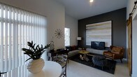 This single-floor space showcases a thoughtfully designed open-concept layout that integrates a living area and dining space into one cohesive environment. The living area is anchored by a striking dark accent wall adorned with a large-scale abstract artwork in warm earth tones of gold, beige, and charcoal, providing a bold focal point. A flat-screen television is mounted on a sleek media console beneath the artwork, complemented by a floor lamp and a plush sectional sofa dressed with accent pillows. Organic-shaped coffee tables in a natural wood finish add texture and warmth to the space.

The dining area features a round white table paired with contemporary chairs, positioned near vertical blinds that filter abundant natural light from floor-adjacent windows. A decorative sunburst wall clock and sculptural accents contribute artistic character throughout. Light-toned flooring runs seamlessly across the space, contrasting effectively with the dark area rug beneath the lounge seating. The high ceiling enhances the sense of openness, while recessed lighting provides ambient illumination, creating a balanced, modern, and inviting atmosphere.