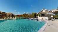 This expansive outdoor pool area is thoughtfully designed to deliver a resort-style experience. The centerpiece is a generously sized swimming pool with crystal-clear turquoise water, complemented by a series of in-water lounge chairs that allow residents to relax partially submerged — a standout feature ideal for warm days. A decorative stacked-stone water feature wall anchors one end of the pool, adding both visual appeal and a soothing ambiance.

The pool deck is finished in a clean, light-toned concrete surface and is surrounded by ample space for sunbathing, with multiple white lounge chairs positioned poolside. A patch of artificial turf adds a touch of greenery alongside the deck area. The adjacent clubhouse structure is constructed with natural stone and board-and-batten siding beneath a distinctive standing-seam metal roof, lending a refined yet relaxed architectural character.

A covered outdoor patio extends from the clubhouse, furnished with dining tables and chairs shaded by striped umbrellas, making it an ideal gathering spot. A grill station is also available, supporting outdoor entertaining. The entire area is enclosed by a wrought-iron perimeter fence, with mature trees providing a scenic natural backdrop under an open, sun-filled sky.
