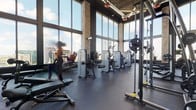 This expansive fitness facility is designed to deliver a comprehensive workout experience across several distinct zones. The main gym area is defined by soaring ceilings, exposed brick columns, and industrial-style ductwork, all paired with walls of floor-to-ceiling windows that frame striking urban skyline views. Rubber flooring throughout this zone supports a generous lineup of strength machines, cable systems, free weights, and treadmills positioned directly against the glass to maximize the cityscape backdrop.

A dedicated cycling studio shares the same architectural character, featuring polished hardwood floors, large mirrors along one wall, and a row of stationary bikes arranged for group sessions. Adjacent to this is a multi-purpose studio with hardwood flooring, a large-screen display, and open wall space adorned with a bold artistic mural, suited for yoga, stretching, or functional training with medicine balls and balance equipment.

An interior transition area features large-format porcelain tile flooring and glass sliding doors that open onto an outdoor terrace, accented with vibrant murals and city views. Together, these zones create a well-equipped, visually dynamic fitness environment elevated high above the surrounding cityscape.