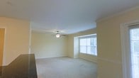 This single-floor space presents a clean and functional living area designed with classic detailing and a warm, neutral palette. The main room features soft, wall-to-wall carpeting in a light beige tone, creating a comfortable and quiet underfoot feel throughout the space. Pale yellow walls contribute to an inviting, airy atmosphere, complemented by crisp white crown molding that runs along the ceiling perimeter, adding a touch of architectural refinement.

A ceiling fan with a built-in light fixture is centrally mounted, providing both ambient lighting and air circulation. Natural light enters through a large, multi-pane window fitted with horizontal blinds, allowing for adjustable light control. The window is framed by a subtle bay-style projection, adding architectural interest and a slight sense of depth to the room.

The space benefits from standard-height ceilings that maintain an open feel without feeling overpowering. White baseboards line the walls throughout, reinforcing the clean, well-maintained aesthetic. Overall, the space offers a straightforward, move-in-ready environment with a cohesive and understated design throughout.