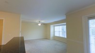 This single-floor space presents a clean and functional living area designed with classic detailing and a warm, neutral palette. The main room features soft, wall-to-wall carpeting in a light beige tone, creating a comfortable and quiet underfoot feel throughout the space. Pale yellow walls contribute to an inviting, airy atmosphere, complemented by crisp white crown molding that runs along the ceiling perimeter, adding a touch of architectural refinement.

A ceiling fan with a built-in light fixture is centrally mounted, providing both ambient lighting and air circulation. Natural light enters through a large, multi-pane window fitted with horizontal blinds, allowing for adjustable light control. The window is framed by a subtle bay-style projection, adding architectural interest and a slight sense of depth to the room.

The space benefits from standard-height ceilings that maintain an open feel without feeling overpowering. White baseboards line the walls throughout, reinforcing the clean, well-maintained aesthetic. Overall, the space offers a straightforward, move-in-ready environment with a cohesive and understated design throughout.