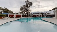 Located at 4550 Atrium Ct in Wilmington, North Carolina, this outdoor amenity space is thoughtfully designed to deliver a resort-like experience. At its centerpiece is a generously sized swimming pool with smooth, curved edges and crystal-clear blue water, complemented by a connected circular spa or wading area. The surrounding pool deck is finished in light-toned concrete, providing a clean, modern aesthetic that reflects the coastal Carolina setting.

Flanking the pool are multiple pergola-covered lounge areas supported by crisp white columns, offering shaded retreats for relaxation. Beneath the pergolas, comfortable seating options include plush daybed-style loungers with gray cushions and sleek white chaise lounges with wood-accented frames, all arranged alongside strips of manicured synthetic turf and stepping stone pathways. Natural river rock detailing at the base of privacy structures adds a refined, organic touch.

Warm, richly grained horizontal wood slat panels serve as privacy screens, creating intimate nooks within the broader communal space. These screens provide visual contrast against the white architectural elements of the surrounding buildings, which feature coastal-inspired shingle and board-and-batten siding with metal roofing. Mature trees and ornamental grasses enhance the natural ambiance, making this a well-appointed outdoor destination.