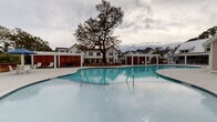 Located at 4550 Atrium Ct in Wilmington, North Carolina, this outdoor amenity space is thoughtfully designed to deliver a resort-like experience. At its centerpiece is a generously sized swimming pool with smooth, curved edges and crystal-clear blue water, complemented by a connected circular spa or wading area. The surrounding pool deck is finished in light-toned concrete, providing a clean, modern aesthetic that reflects the coastal Carolina setting.

Flanking the pool are multiple pergola-covered lounge areas supported by crisp white columns, offering shaded retreats for relaxation. Beneath the pergolas, comfortable seating options include plush daybed-style loungers with gray cushions and sleek white chaise lounges with wood-accented frames, all arranged alongside strips of manicured synthetic turf and stepping stone pathways. Natural river rock detailing at the base of privacy structures adds a refined, organic touch.

Warm, richly grained horizontal wood slat panels serve as privacy screens, creating intimate nooks within the broader communal space. These screens provide visual contrast against the white architectural elements of the surrounding buildings, which feature coastal-inspired shingle and board-and-batten siding with metal roofing. Mature trees and ornamental grasses enhance the natural ambiance, making this a well-appointed outdoor destination.