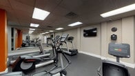 The fitness center at River Oak Apartments is a comprehensive, single-floor amenity space designed to support a full range of workout routines. The facility is organized across several interconnected areas, each dedicated to a specific type of training. Flooring throughout the gym consists of durable rubber matting, providing traction and impact absorption suitable for high-intensity use.

The cardio area is equipped with multiple treadmills and elliptical machines by Life Fitness, arranged in rows alongside full-length wall mirrors that enhance the sense of space. A wall-mounted flat-screen television is positioned for easy viewing during workouts. Recessed panel lighting in a drop ceiling provides consistent, even illumination throughout.

A separate strength training area houses a multi-station cable machine, a dedicated bench press with barbell and weight plates, a dumbbell rack, and additional resistance equipment. An accent wall in a bold orange hue adds a distinctive visual element to this zone. A water fountain is conveniently mounted nearby.

The facility also includes a full racquetball court featuring a polished hardwood floor with red boundary lines, white panel walls, and overhead lighting — a standout feature that sets this fitness center apart from typical community gyms.