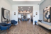 Located at 26120 Jones Loop Rd in Punta Gorda, Florida, this thoughtfully designed single-story community space offers a generous, open-concept layout that seamlessly blends a fully appointed kitchen with an inviting living area. The entryway makes an immediate impression with its distinctive geometric-patterned hardwood flooring, transitioning into wide-plank wood-look flooring that flows throughout the entire space.

The kitchen features an expansive white quartz island with navy blue cabinetry, illuminated by elegant glass pendant lights with brass accents. Open shelving with blue tile backsplash provides both style and function, while shiplap accent walls add coastal charm. A coffered ceiling with exposed wood beams and a chandelier defines the dining and gathering zone, lending architectural character to the open floor plan.

The living area centers around a striking floor-to-ceiling patterned tile fireplace wall, complemented by large windows dressed in sheer white curtains that flood the room with natural light. A dedicated workspace nook features a built-in desk set against a shiplap accent wall, adorned with curated modern artwork. Throughout, carefully selected blue-toned furniture, contemporary art pieces, and thoughtful lighting create a cohesive, welcoming coastal-modern ambiance.