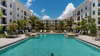 This striking multi-story residential complex showcases a thoughtfully designed outdoor amenity space that serves as its centerpiece. The property features a contemporary white stucco exterior with clean architectural lines, large windows, and private balconies that overlook the vibrant courtyard below. The complex spans two floors and is arranged in a U-shaped or courtyard configuration, allowing residents on multiple levels to enjoy sweeping views of the resort-style grounds.

At the heart of the outdoor area lies a generously sized swimming pool with crystal-clear blue water, complemented by a second pool area visible across the landscaped grounds. The expansive pool deck is finished with elegant stone pavers and furnished with an abundance of lounge chairs, providing ample space for relaxation and sunbathing. Raised planting beds with tropical foliage and mature palm trees are thoughtfully integrated throughout the deck, adding to the lush, resort-like atmosphere.

Ornamental iron fencing defines the perimeter of the pool areas, while additional green lawn spaces and shade structures — including pergolas and umbrella stations — offer comfortable retreats from the Florida sun. The tropical landscaping, featuring tall palm trees and manicured plantings, reinforces the warm, relaxed ambiance of the complex. The entire outdoor setting benefits from abundant natural light and open skies, creating an inviting, vacation-inspired environment.