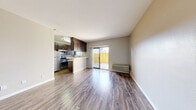This single-floor unit offers a well-organized layout that maximizes its compact footprint. The main living area is anchored by warm wood-look flooring that runs throughout the space, neutral-toned walls, and a ceiling fan that adds both function and a finishing touch. A sliding glass door floods the room with natural light and provides direct access to a private outdoor patio enclosed by a yellow fence, offering a pleasant outdoor extension of the living space. A wall-mounted HVAC unit ensures climate control within the main area.

The kitchen is positioned adjacent to the living space in an open-concept arrangement, featuring dark espresso cabinetry extending to the ceiling for ample storage, a mosaic tile backsplash, white countertops, and stainless steel appliances including a refrigerator, gas range, and microwave. A peninsula counter with an overhang separates the kitchen from the living area, providing additional workspace or casual seating.

The bathroom is well-appointed with a dark wood vanity, white countertop, a large mirror with overhead lighting, and wood-look flooring. A dedicated closet provides convenient storage. Overall, the space presents a clean, modern aesthetic with practical finishes throughout.