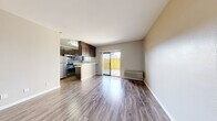 This single-floor unit offers a well-organized layout that maximizes its compact footprint. The main living area is anchored by warm wood-look flooring that runs throughout the space, neutral-toned walls, and a ceiling fan that adds both function and a finishing touch. A sliding glass door floods the room with natural light and provides direct access to a private outdoor patio enclosed by a yellow fence, offering a pleasant outdoor extension of the living space. A wall-mounted HVAC unit ensures climate control within the main area.

The kitchen is positioned adjacent to the living space in an open-concept arrangement, featuring dark espresso cabinetry extending to the ceiling for ample storage, a mosaic tile backsplash, white countertops, and stainless steel appliances including a refrigerator, gas range, and microwave. A peninsula counter with an overhang separates the kitchen from the living area, providing additional workspace or casual seating.

The bathroom is well-appointed with a dark wood vanity, white countertop, a large mirror with overhead lighting, and wood-look flooring. A dedicated closet provides convenient storage. Overall, the space presents a clean, modern aesthetic with practical finishes throughout.