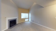 This two-floor space showcases a thoughtfully designed living area as its centerpiece. The room immediately draws attention with its vaulted ceiling, which rises dramatically to create an open, airy atmosphere. Neutral gray carpet covers the floor, complementing the light gray and soft yellow walls that lend the space a warm yet understated tone.

A tile-surround fireplace with a classic white mantel anchors one wall, serving as a functional and decorative focal point. Opposite the fireplace, a large window paired with a glass-paneled door floods the room with natural light, offering views of the surrounding exterior. Horizontal blinds on both the window and door provide privacy and light control as needed.

The angled ceiling design adds architectural character, with the vaulted section transitioning to a standard ceiling height toward the rear of the room. White baseboards frame the perimeter cleanly, and a floor-level air vent indicates a built-in HVAC system. The overall ambiance is clean, open, and move-in ready, with a layout that allows for flexible furniture arrangement across the spacious living area.