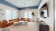 Located at 12609 Matisse Cir in New Port Richey, Florida, this single-floor community space showcases a thoughtfully designed interior with a distinct coastal aesthetic. The layout is divided into two primary areas that flow seamlessly together, unified by a calming blue and white color scheme with crisp white walls and rich carpeted flooring throughout.

The lounge area is a welcoming, generously sized gathering space anchored by a large wraparound leather sectional sofa in warm cognac tones. Recessed lighting and elevated ceilings with a bold blue upper accent band add dimension and visual interest to the room. A large flat-screen television mounted above a sleek dark media console serves as a focal point, while layered coffee tables and upholstered ottomans complete the relaxed, functional layout. Lush potted fig trees and curated wall art lend a fresh, lived-in atmosphere.

The dining and bar area features a light wood communal table surrounded by contemporary gray upholstered chairs, complemented by a white subway tile accent wall adorned with coastal-themed framed artwork. Industrial-style gooseneck sconce lighting illuminates a counter-height bar lined with natural wood stools. A beverage refrigerator and utility sink are integrated into the cabinetry, making this space well-equipped for social gatherings. Together, these areas create an inviting, resort-inspired environment ideal for community engagement.