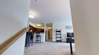 This single-floor space is designed with an open-concept layout that seamlessly connects the living area, dining space, and kitchen. The living area features light-colored carpeting and neutral white walls, creating a clean, understated backdrop. A lean ladder-style shelving unit and a flat-screen television on a compact media stand anchor the space, while a framed piece of wall art adds a personal touch.

The kitchen is positioned toward the back left of the main area and features dark wood cabinetry that contrasts with the lighter surroundings. A raised breakfast bar with bar-height seating provides a casual dining option, making the kitchen both functional and social. Ceiling-mounted lighting illuminates the kitchen area effectively.

A hallway visible from the main living space leads to additional rooms, suggesting a practical and well-organized floor plan. The transition between spaces flows naturally, and the consistent use of neutral tones throughout contributes to a cohesive and welcoming ambiance. Overall, the space is functional and comfortable, suited for everyday living.