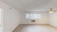 This single-floor unit offers a clean, modern aesthetic throughout, characterized by a consistent palette of crisp white walls, light wood-style flooring, and textured ceilings. The living area is open and airy, anchored by a wide horizontal window fitted with vertical blinds that frames views of lush greenery outside, along with a wall-mounted air conditioning unit for year-round comfort. The standout feature of the unit is the galley-style kitchen, which has been thoughtfully renovated with bold navy blue shaker cabinetry, brushed gold hardware, and striking white quartz countertops with subtle veining. Stainless steel appliances, including a refrigerator, electric range, and microwave, complete this well-appointed cooking space, while tile flooring ties the design together. The bedrooms are quiet and well-lit, each featuring windows with vertical blinds that allow natural light to filter in. The bathroom is finished with a marble-look quartz vanity countertop, dual mirrors, brushed nickel fixtures, and a fully tiled walk-in shower equipped with a rain-style showerhead and a built-in bench seat. Throughout the unit, clean lines, neutral tones, and updated finishes create a cohesive and functional living environment.