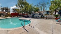 The outdoor amenity area at Avenida Alcachofa in San Diego's 92128 zip code centers around a generously sized freeform swimming pool with clear turquoise water, complemented by a separate spa with stainless steel entry rails. The expansive concrete pool deck provides ample space for lounge seating and is furnished with multiple chairs and umbrella-shaded dining sets in vibrant orange and red tones, ideal for relaxing in San Diego's sunny climate. Tall palm trees and mature deciduous trees frame the perimeter, offering natural shade and a lush, resort-like atmosphere. A distinctive pergola structure with textured stucco columns anchors a dedicated seating area, providing additional covered outdoor space with picnic-style tables and colorful accent chairs beneath a slatted overhead canopy that filters sunlight. The pool facility building features a stucco exterior consistent with the surrounding Spanish-influenced residential architecture, complete with red-tiled rooflines visible in the background. Raised planter beds with drought-tolerant succulents and native plants add a decorative, low-maintenance landscaping element near the facility entrance. The overall environment is well-maintained and designed to encourage outdoor social activity year-round.