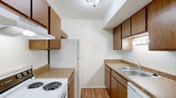 This single-floor unit offers a straightforward, functional layout designed for comfortable everyday living. The galley-style kitchen is efficiently organized, featuring warm wood-toned cabinetry on both upper and lower levels, granite-look laminate countertops, and wood-look vinyl flooring. White appliances, including a coil-burner electric range with overhead ventilation, a full-size refrigerator, and a dishwasher, are neatly integrated alongside a double-basin stainless steel sink. The open living area transitions from wood-look vinyl flooring to plush neutral carpet, creating a clear distinction between functional and relaxing zones. A large window fitted with vertical blinds allows natural light to fill the room, while a wall-mounted thermostat and a window AC unit provide climate control. The bedroom is carpeted and includes a built-in closet with a wood-paneled sliding door, offering practical storage. The bathroom is clean and simply appointed, featuring wood-look vinyl flooring, a full-size soaking tub with a tub/shower surround, a toilet, and a wood-toned vanity with a granite-look countertop. Throughout the unit, white walls and textured ceilings maintain a bright, neutral aesthetic.