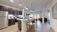This single-story, one-bedroom, one-bathroom unit offers a thoughtfully designed open-concept layout that maximizes its compact footprint. Light wood-style flooring flows throughout the space, lending warmth and visual continuity across every room.

The kitchen and living area form the heart of the unit. The kitchen is equipped with stainless steel appliances, including a refrigerator, electric range, and microwave, all complemented by rich dark-espresso cabinetry extending to the ceiling for ample storage. A granite-topped island with seating for two serves as both a functional workspace and casual dining area, illuminated by stylish pendant lighting. Recessed ceiling lights and under-cabinet lighting further enhance the kitchen's functionality.

The living area connects seamlessly to the kitchen and features a ceiling fan and direct access to an outdoor balcony through French-style doors, bringing in natural light and outdoor views. The bedroom is a comfortable, well-proportioned retreat with a large window overlooking greenery and a ceiling fan overhead.

The bathroom features a granite-topped dark vanity and wood-style flooring, with direct access to a dedicated closet with built-in hanging rods. A separate in-unit laundry area includes a stacked Whirlpool washer and dryer alongside wire shelving for additional storage.