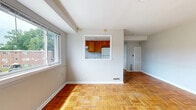 Located in Baltimore, MD 21218, this well-laid-out one-bedroom, one-bathroom unit offers a functional and comfortable living environment across a single floor. The living room is a welcoming, open space highlighted by warm parquet hardwood flooring in a distinctive basket-weave pattern and a large window that draws in natural light and outdoor views. A charming pass-through window connects the living area to the kitchen, which features warm wood cabinetry, a white refrigerator, and a bright, efficient layout. The hallway, accessible from the living room, leads to the remaining rooms and includes a louvered closet door for additional storage. The bedroom is a generously sized retreat with smooth hardwood floors, a wide window overlooking a green outdoor setting, and a full double-door closet providing ample storage space. The bathroom is finished with clean white subway tile walls, tile flooring, and a combined shower and tub enclosed by sliding glass doors, complemented by a small window for natural ventilation. Neutral gray walls throughout the unit create a cohesive, move-in-ready aesthetic. The overall layout is practical and efficient, making the most of the available space.