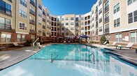 Located at 501 Willard St in Durham, North Carolina, this beautifully appointed courtyard amenity space is nestled within a multi-story residential community and designed to offer a resort-like outdoor experience. At its centerpiece is a generously sized swimming pool with crystal-clear blue water, framed by a wide concrete and wood-plank sundeck outfitted with lounge chairs and shaded cabanas — ideal for relaxing under the North Carolina sun.

The surrounding deck transitions seamlessly into a warm, richly toned wooden terrace that serves as an outdoor gathering hub. Here, residents will find multiple seating arrangements shaded by large patio umbrellas, alongside a stone-accented outdoor fireplace that adds a cozy, communal focal point. A well-equipped outdoor kitchen with a stainless steel grill is also integrated into the space, perfect for al fresco dining and entertaining.

Lush landscaping with manicured greenery, raised planters, and decorative lighting strung overhead enhance the ambiance throughout. The entire courtyard is embraced by the community's multi-story brick and siding exterior with private balconies overlooking the space, creating an intimate, enclosed atmosphere. The thoughtful design balances active recreation with relaxed social spaces, making this courtyard a standout amenity within the Durham community.