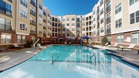 Located at 501 Willard St in Durham, North Carolina, this beautifully appointed courtyard amenity space is nestled within a multi-story residential community and designed to offer a resort-like outdoor experience. At its centerpiece is a generously sized swimming pool with crystal-clear blue water, framed by a wide concrete and wood-plank sundeck outfitted with lounge chairs and shaded cabanas — ideal for relaxing under the North Carolina sun.

The surrounding deck transitions seamlessly into a warm, richly toned wooden terrace that serves as an outdoor gathering hub. Here, residents will find multiple seating arrangements shaded by large patio umbrellas, alongside a stone-accented outdoor fireplace that adds a cozy, communal focal point. A well-equipped outdoor kitchen with a stainless steel grill is also integrated into the space, perfect for al fresco dining and entertaining.

Lush landscaping with manicured greenery, raised planters, and decorative lighting strung overhead enhance the ambiance throughout. The entire courtyard is embraced by the community's multi-story brick and siding exterior with private balconies overlooking the space, creating an intimate, enclosed atmosphere. The thoughtful design balances active recreation with relaxed social spaces, making this courtyard a standout amenity within the Durham community.