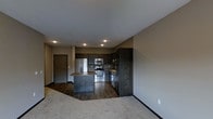 This single-floor space showcases an open-concept layout that integrates the kitchen and living area into one cohesive environment. The kitchen is outfitted with rich, dark wood cabinetry that extends from the lower counters to the upper cabinets, providing ample storage throughout. A central island with a neutral-toned countertop serves as both a workspace and a natural dividing point between the kitchen and living area. Stainless steel appliances, including a refrigerator, microwave, and range, lend a modern, polished look to the cooking space.

The flooring transitions from dark hardwood in the kitchen area to plush carpeting in the living space, subtly defining each zone while maintaining an open feel. Recessed lighting is installed throughout the ceiling, providing even, practical illumination across the space. The neutral gray and taupe wall tones complement the dark cabinetry and flooring, creating a cohesive and contemporary aesthetic. A single entry door is visible along one wall, and the overall layout feels efficient and well-organized, making practical use of the available footprint.